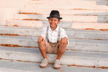 Cute fashionable African-American boy sitting on stairs outdoors�