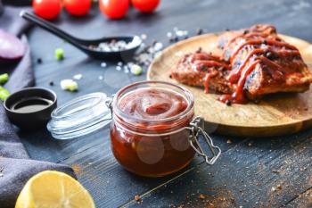 Jar with barbecue sauce and fried meat on dark table�