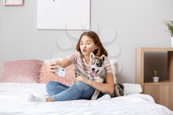 Cute teenage girl with funny husky puppy taking selfie on bed at home�