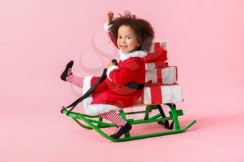 Little African-American girl in Santa costume, with sledges and gift boxes on color background�