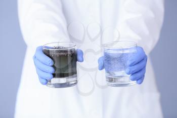 Female doctor with glasses of dirty and clean water on color background, closeup�
