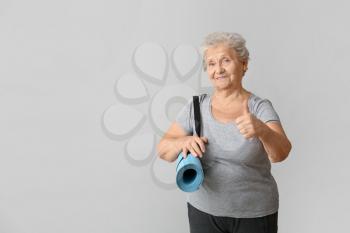 Elderly woman with yoga mat showing thumb-up gesture on light background�