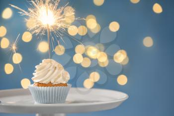 Delicious birthday cupcake with sparkler on stand against blurred lights�