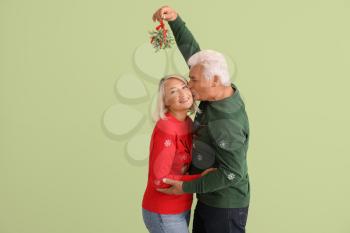 Mature man kissing his wife under mistletoe branch on color background�