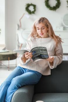 Beautiful young woman in warm sweater reading magazine at home�