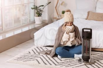 Young woman in warm clothes and with cup of tea sitting near electric heater at home. Concept of heating season�