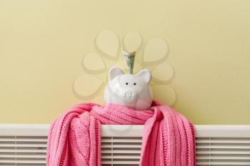 Piggy bank with money and scarf on radiator. Concept of heating season�