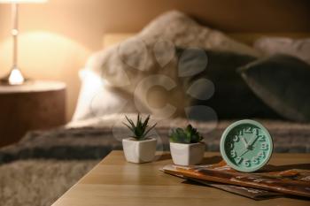 Alarm clock with magazines and houseplants on table in bedroom at night�