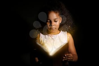 Little African-American girl reading magic book on dark background�