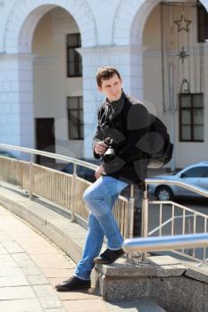Young man in black coat and blue jeans posing with camera outdoors
