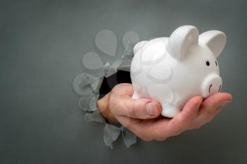 Hand with a piggy bank through a grey paper hole. Financial concept.