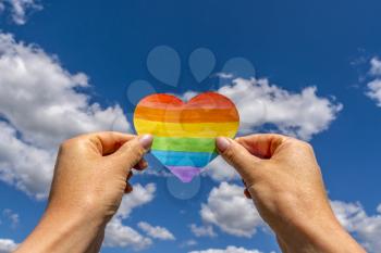 LGBT rights concept, hands holds a heart painted like a LGBT flag against sky background