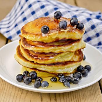 A stack of pancakes with blueberries and honey on a white plate, napkin against a wooden board