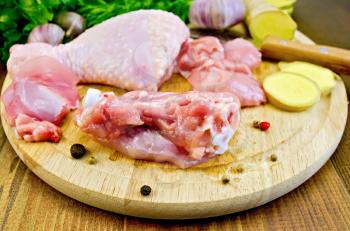 Chicken leg cut on a round board, garlic, parsley, ginger, dill, pepper and a knife on a wooden boards background