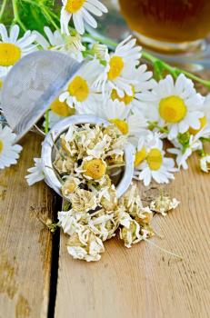 Metal strainer with dried chamomile, a bouquet of fresh flowers chamomile, tea in a glass cup on a background of wooden boards
