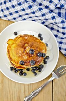 A stack of pancakes with blueberries and honey on a white plate, napkin, fork on the background of wooden boards on top