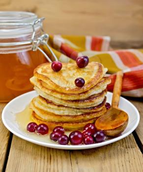 A stack of pancakes with cranberries and honey, wooden spoon on a white plate, a jar of honey, a napkin on a wooden boards background