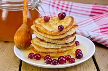 A stack of pancakes with cranberries and honey on a white plate with honey jar, a wooden spoon, napkin against a wooden board