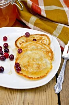 Pancakes with cranberries and honey on a white plate, a jar of honey, fork, napkin on the background of wooden boards