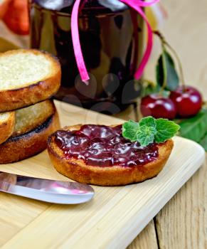 Slices of toasted bread, a glass jar with cherry jam, knife on background wooden board