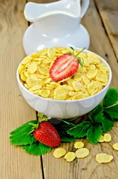 Cornflakes in a white bowl, leaves and strawberries, milk in a jug on a wooden boards background
