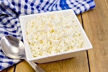 Cottage cheese in a white square bowl, blue checkered napkin, spoon on a wooden board