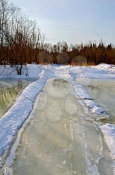 Melting ice on the river, melted road alternating river, snow, bushes and trees on the shore against the blue sky
