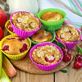 Cupcakes with rhubarb, cherries, apples in tins, milk in a jug, a napkin on the background of wooden boards