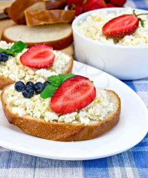 Slices of bread with curd cream, blueberries and strawberries, mint, a bowl of cottage cheese, bread on a board on a background of the blue checkered tablecloth