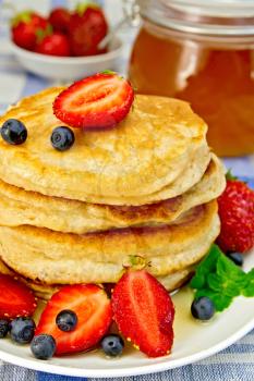 A stack of pancakes with strawberries, blueberries and honey on a white plate, a jar of honey on a linen tablecloth background