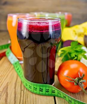 Beet juice in the foreground and juice from carrot, pumpkin, cucumber and tomato in a tall glass, vegetables on background of wooden boards