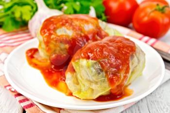 Stuffed cabbage meat in cabbage leaves with tomato sauce in the plate on a napkin, tomatoes, parsley and garlic on a background of pale wooden plank