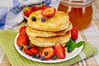 A stack of pancakes with strawberries, blueberries and honey on a white plate, jars of honey, napkins on a background of linen tablecloths