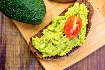 A slice of rye bread with guakomole and tomatoes, avocado on a wooden boards background