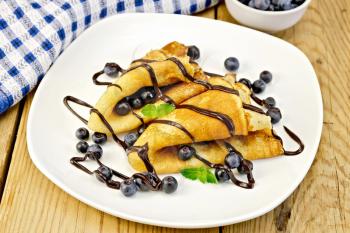 Two pancakes with blueberries, mint and chocolate icing on a plate, napkin, bowl on a wooden boards background