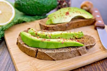 Two slices of rye bread with slices of avocado and pepper, lemon, napkin against the dark wooden board