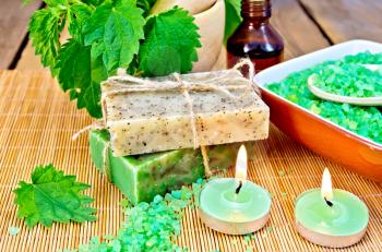 Two bars of homemade soap with twine, bath salt in a bowl, oil in the bottle, nettles in a mortar, two candles on a wooden boards background