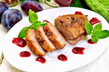 Roasted duck breast with plum sauce and basil in a white oval plate, towel and plums on a background of light wooden boards
