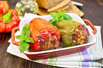 Two sweet peppers stuffed with meat and rice with basil leaves in a brown roasting pan on a napkin on a dark wooden boards background