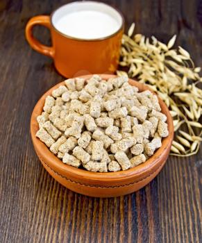 Oat bran large in earthenware bowl, a mug with milk, oat stalks on a wooden boards background