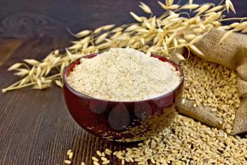 Oat flour in a bowl, bag with oat and stalks of oats on the background of dark wood planks