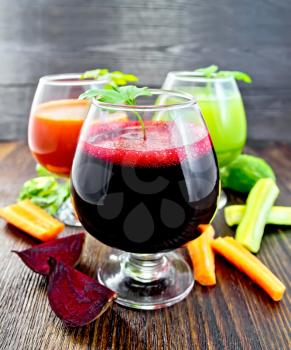 The juice of beet, carrot and cucumber in three wineglass, vegetables and parsley on a wooden board background