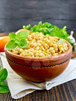 Barley porridge in a clay bowl with basil on a kitchen towel, parsley and garlic on a wooden boards background