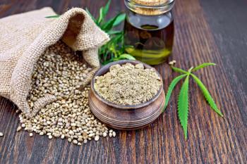 Hemp flour in a clay bowl, the grain in the bag and on the table, the oil in a glass jar, leaves and stalks of cannabis on a background of dark wooden boards