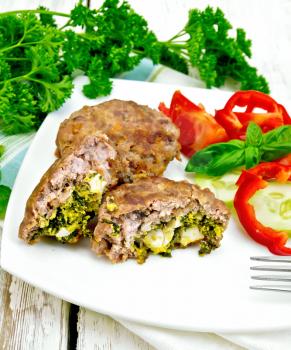 Cutlets stuffed with spinach and egg, salad with tomatoes, cucumber and pepper in a dish on a napkin, basil and parsley on the background light wooden boards