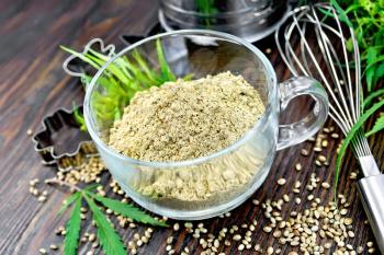 Hemp flour in a glass cup, mixer, sieve and molds for cookies, green leaves and cannabis seeds on the background of dark wood planks