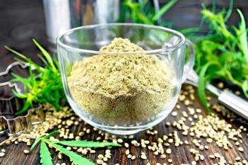 Hemp flour in a glass cup, mixer, sieve and molds for cookies, green leaves and cannabis seeds on the background of wooden boards