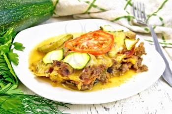 Casserole of minced meat, tomato and zucchini in a plate, napkin and fork on a light wooden board background