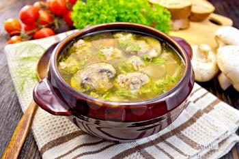 Soup with meatballs, noodles and champignon in a clay bowl on a napkin, parsley, tomatoes, mushrooms and bread on a wooden boards background