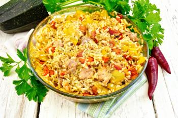 Rice with chicken, tomatoes, carrots, zucchini and garlic in a glass pan on a napkin, parsley and hot pepper on the background light wooden boards